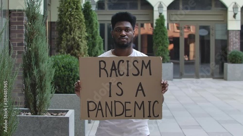 A young African American man stands with a cardboard poster RACISM IS A PANDEMIC in a public outdoor place. Street demonstration. Slow motion. Close up. photo