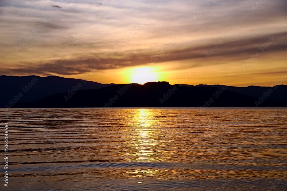 夕暮れの湖。日暮れの太陽に黄金色に輝く湖面。周囲の山のシルエット。