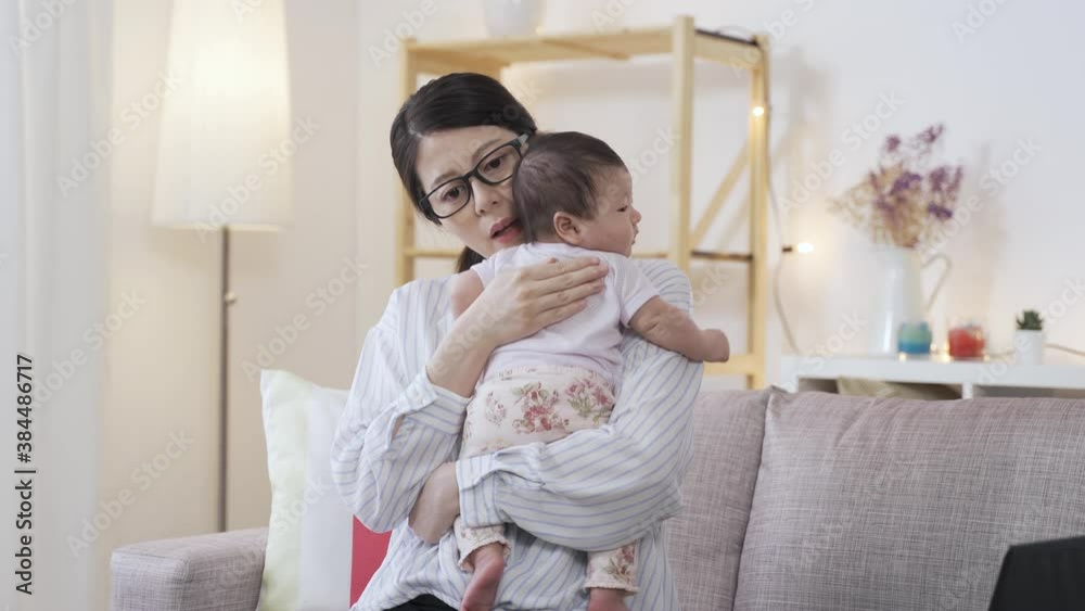 portrait chinese working mother is burping her adorable baby by patting on its back on the sofa in a cozy living room. portrait chinese working mother is burping her adorable baby by patting on its back on the sofa in a cozy living room.