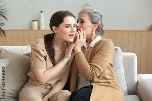 Happy middle mother whispering secret to her smiling daughter at home, gossiping, sharing secret. Beautiful woman hearing good news from mature mum, trusted relations.