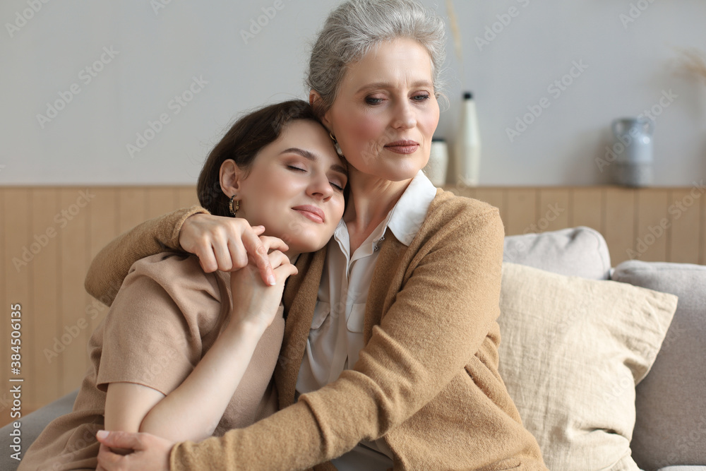 Cheerful young woman is embracing her middle aged mother with closed eyes hugging, touching cheeks, sitting on couch at home. Happy trusted relations. Family concept. Cheerful young woman is embracing her middle aged mother with closed eyes hugging, touching cheeks, sitting on couch at home. Happy trusted relations. Family concept.
