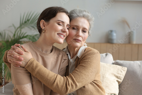 Cheerful young woman is embracing her middle aged mother with closed eyes hugging, touching cheeks, sitting on couch at home. Happy trusted relations. Family concept.