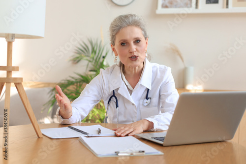 Older woman doctor therapist wearing headset video call talking to web camera consulting virtual patient online by video conference call chat.