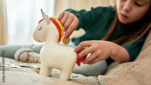 A little girl holding a red tail of her porcelian unicorn moneybox and putting a coin into a slot while sitting on a sofa in a big room photo