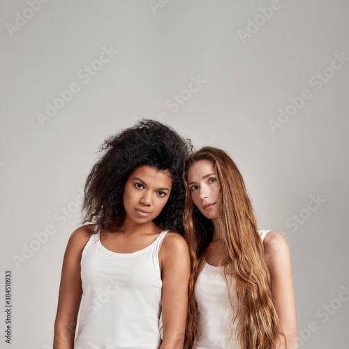 Portrait of two young diverse women wearing white shirts looking at camera while standing together isolated over grey background