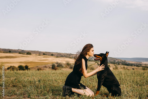 Young woman sitting on green grass, hugging a doberman breed dog, touching noses. © Bostan Natalia