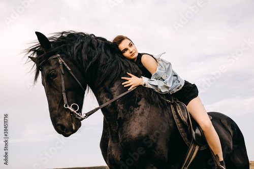 Young woman wearing a denim jacket, riding a black horse, outdoors.