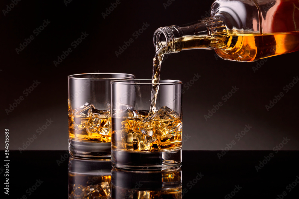 Pouring whiskey from bottle to two glasses with ice cubes on black background.