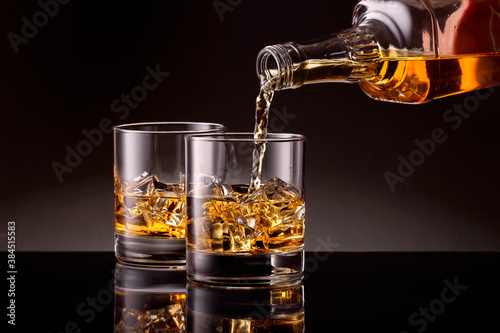 Obraz na płótnie Pouring whiskey from bottle to two glasses with ice cubes on black background. w salonie