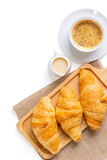 Croissants on wooden bread cutting board with sackcloth and with sweetened condensed milk, cappuccino coffee mug, over white background. Croissant french breakfast. Top view.