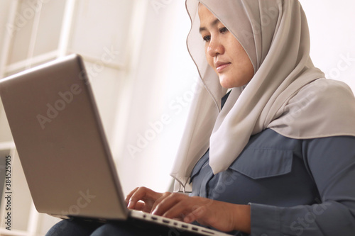 Asian muslim woman wearing hijab smiling while typing on her laptop, work from home concept. Young successful female entrepreneur working at home