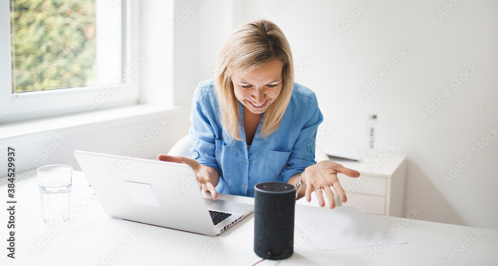 beautiful young and blond woman is talking to her smart speaker and is working at home during corona lock down beautiful young and blond woman is talking to her smart speaker and is working at home during corona lock down