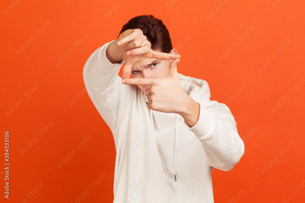 Concentrated creative young man in casual hoodie rising hands to eyes and making frame looking for suitable shot, cameraman job. Indoor studio shot isolated on orange background Concentrated creative young man in casual hoodie rising hands to eyes and making frame looking for suitable shot, cameraman job. Indoor studio shot isolated on orange background