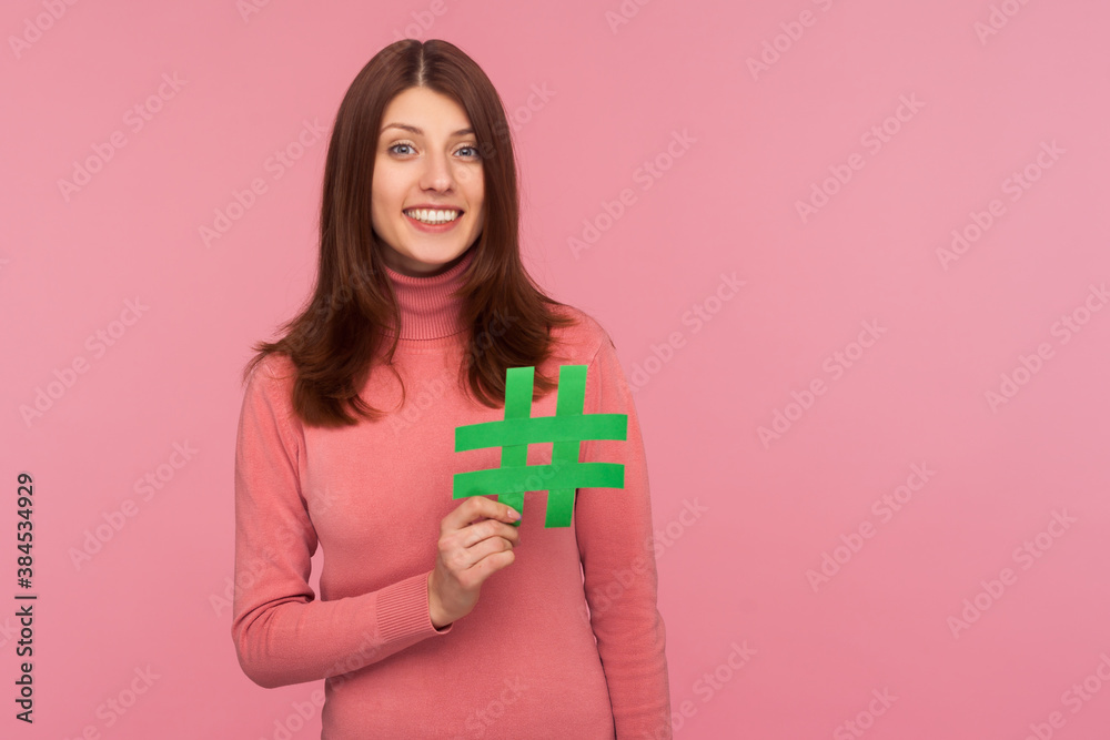 Happy brunette woman in pink sweater holding green hashtag sign looking at camera with toothy smile, easy blog search. Indoor studio shot isolated on pink background Happy brunette woman in pink sweater holding green hashtag sign looking at camera with toothy smile, easy blog search. Indoor studio shot isolated on pink background