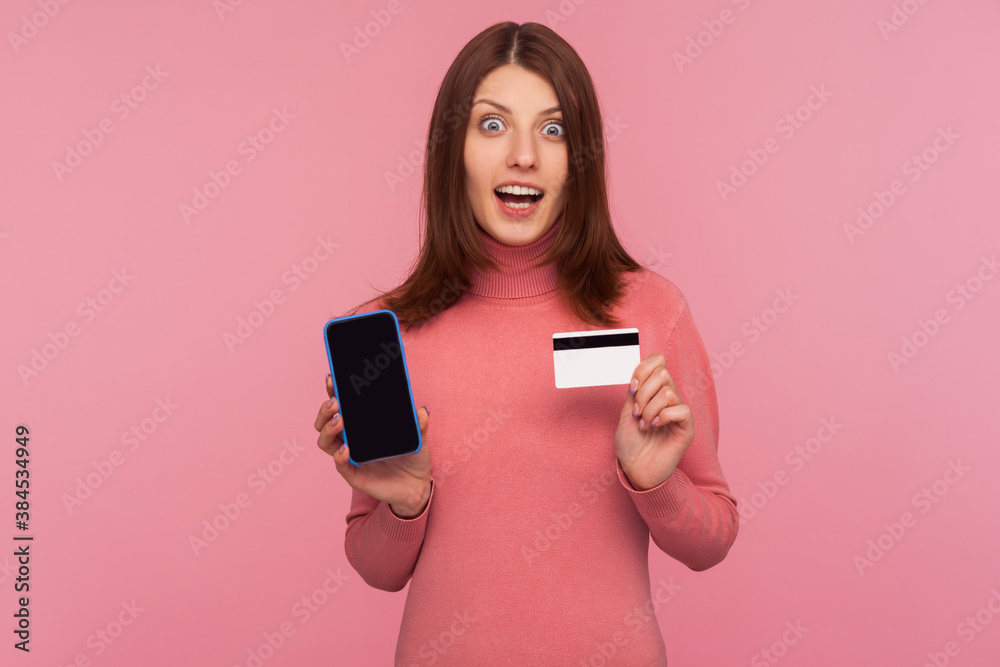 Amazed brunette woman in pink sweater showing cell phone and credit card, looking at camera with shocked expression, surprized with online banking. Indoor studio shot isolated on pink background Amazed brunette woman in pink sweater showing cell phone and credit card, looking at camera with shocked expression, surprized with online banking. Indoor studio shot isolated on pink background