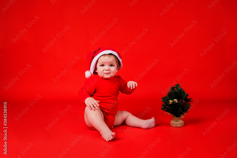 playful baby girl in a red bodysuit in a red Christmas hat sitting with a Christmas tree on a red background playful baby girl in a red bodysuit in a red Christmas hat sitting with a Christmas tree on a red background