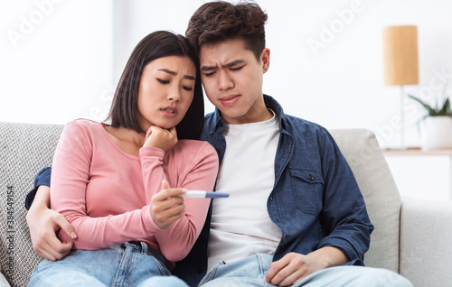 Sad Asian Couple Holding Negative Pregnancy Test Sitting At Home