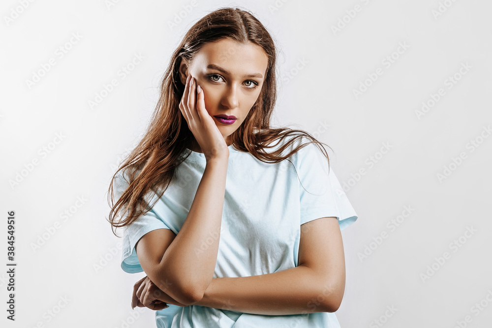 Beautiful young brunette girl with green eyes with long hair with fashion makeup with purple lips on an isolated gray background. The woman holds hand to face and seriously looking at the camera