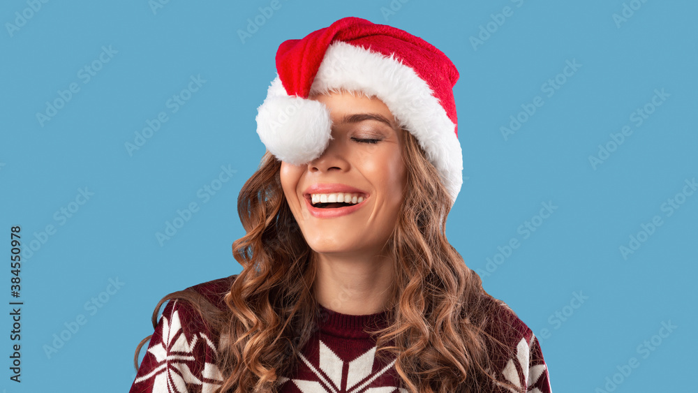 Portrait of silly young lady in warm clothes having fun with Santa hat on blue studio background, panorama