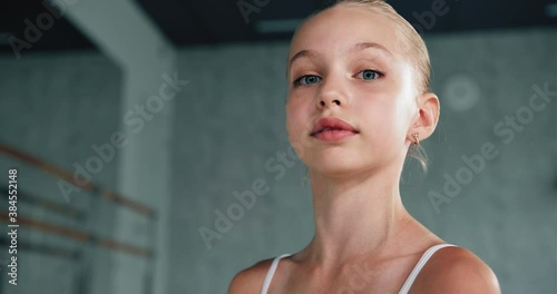 Pretty blonde girl dancer with blue eyes in white suit with hair bun on head poses for shoot as actress in ballet studio closeup