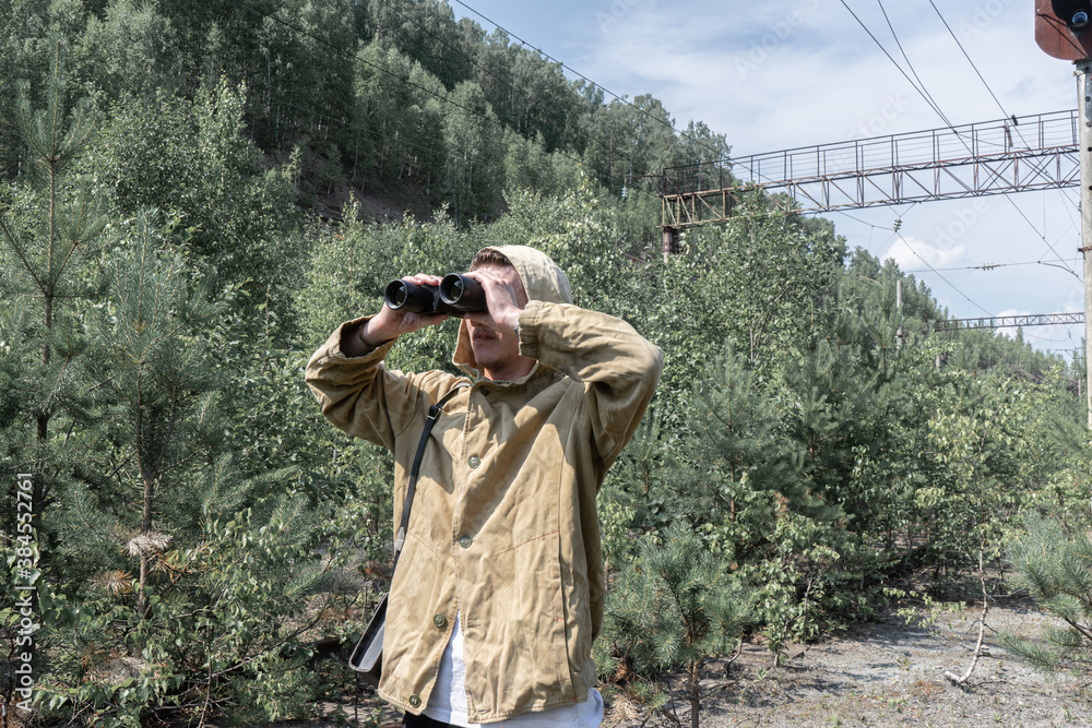 a young man stands in front of a railway traffic light and looks through binoculars. abandoned railway in the forest.