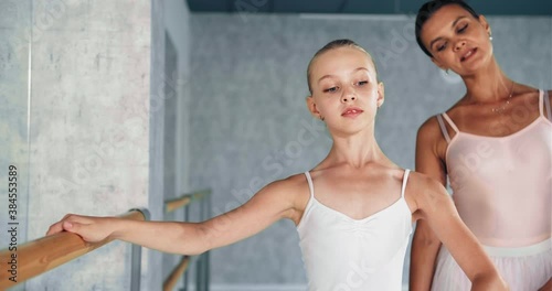 Female trainer helps preteen ballerina do dance exercises leaning on wooden bar near large mirror in studio at dance lesson photo