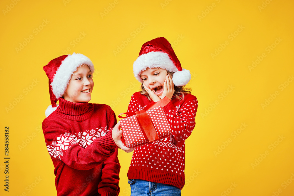 Fototapeta premium Happy boy in Santa hat giving Christmas gift box to excited girl in red sweater with deer on yellow background