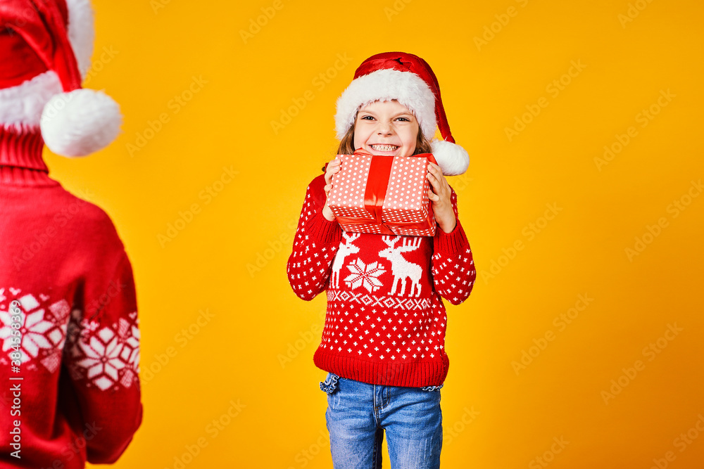 Fototapeta premium Happy boy in Santa hat giving Christmas gift box to excited girl in red sweater with deer on yellow background