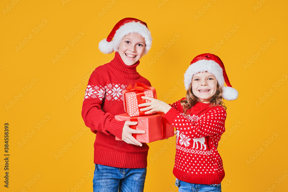 Cheerful kids in Santa hats and red sweaters standing with presents laughing and looking at camera on yellow background Cheerful kids in Santa hats and red sweaters standing with presents laughing and looking at camera on yellow background