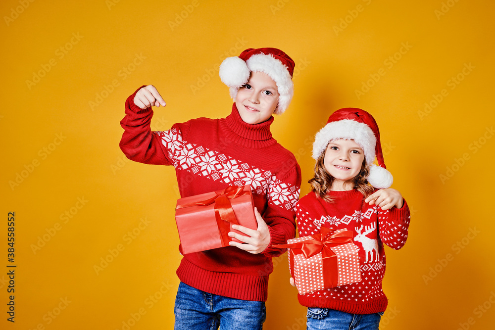 Fototapeta premium Cheerful kids in Santa hats and red sweaters standing with presents laughing and looking at camera on yellow background