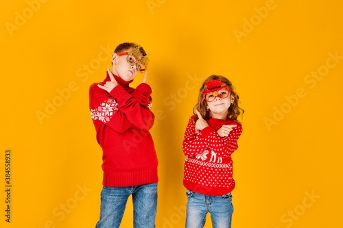 Children in warm red Christmas sweaters and decorated glasses looking at camera on yellow background.