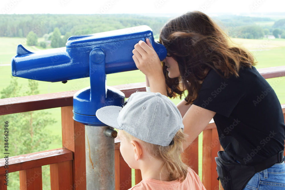 Teenage girl and little brother looking through blue tourist telescope or sightseeing monocular explore green landscape.