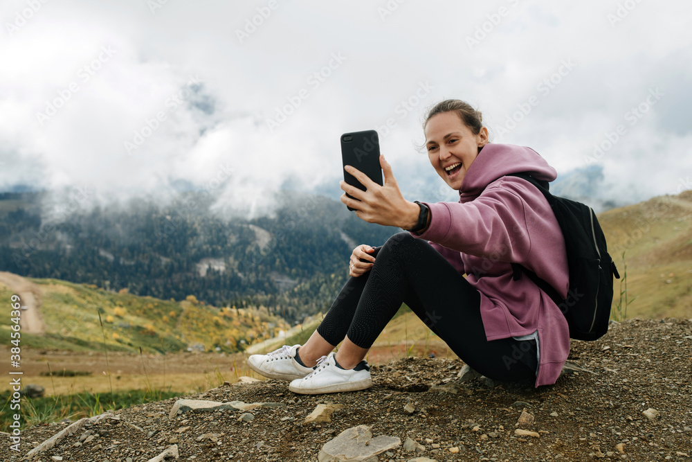 Cheerful woman taking selfie, sitting on the ground high up in cloudy mountains