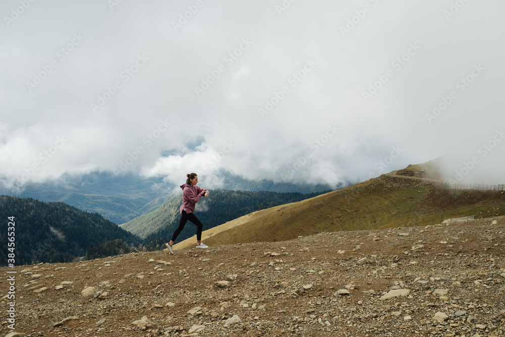 Far away shot of young woman running high up in cloudy mountains. Side view.