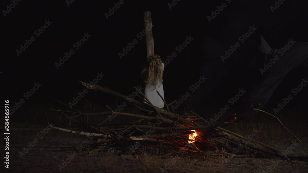 Hand of inquisitor in black shroud igniting bonfire with flaming torch with innocent hopeless woman in white dress tied to wooden pole, convicted as witch, outdoors at night during medieval witchhunt.