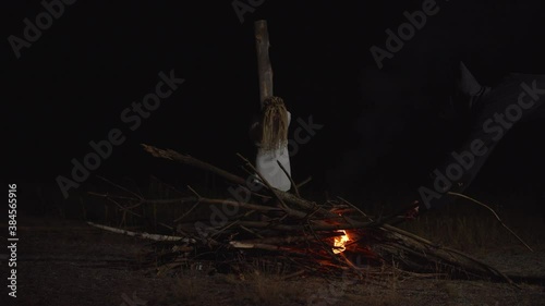 Hand of inquisitor in black shroud igniting bonfire with flaming torch with innocent hopeless woman in white dress tied to wooden pole, convicted as witch, outdoors at night during medieval witchhunt.