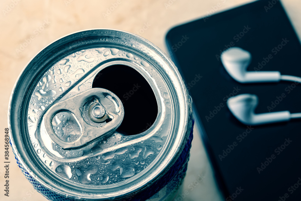 Open cold beer can with water drops near smartphone and earphones. Drinking on the go beverages in aluminum cans with pull tabs. Open cold beer can with water drops near smartphone and earphones. Drinking on the go beverages in aluminum cans with pull tabs.