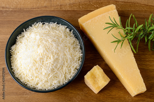 Grated Parmesan or Grana Padano in a ceramic bowl, wedge of hard cheese and rosemary on a brown cutting board. Delicious ingredient for pizza, sandwiches, salads.