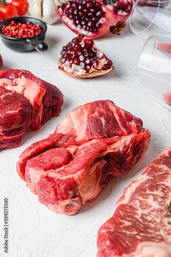 Beef cuts Chuck eye roll, top blade, rump steak with red wine in glass and bottle, herbs and pomegranate. Organic meat. White textured background. Side view vertical.