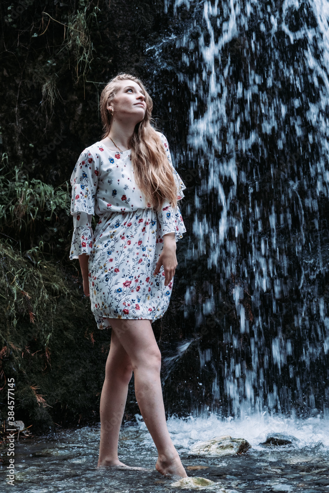 Vertical shot of an attractive young woman in a light summer dress posing with a waterfall behind Vertical shot of an attractive young woman in a light summer dress posing with a waterfall behind