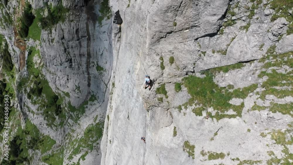 Aerial shot of two people climbing up a very steep cliff on a mountain together in the alps of Switzerland during a sunny summer day. Green grass beneath them as they climb up on a rope ladder. Aerial shot of two people climbing up a very steep cliff on a mountain together in the alps of Switzerland during a sunny summer day. Green grass beneath them as they climb up on a rope ladder.