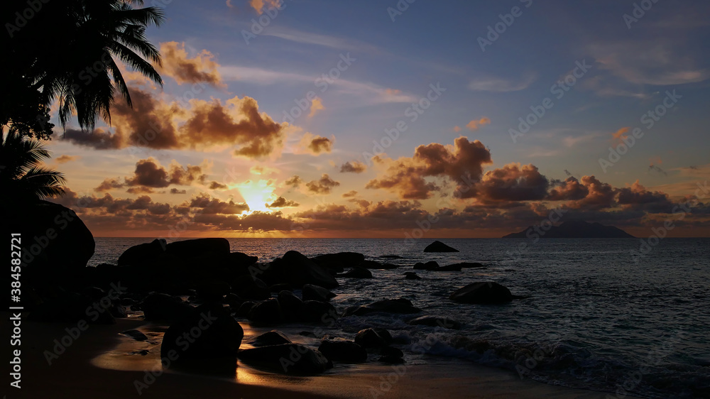 Majestic sunset on tropical Tusculum Beach, Mahe island, Seychelles with sun covered by clouds over horizon and visible sunbeams. Silhouettes of coconut palm and granite rock formations in foreground.