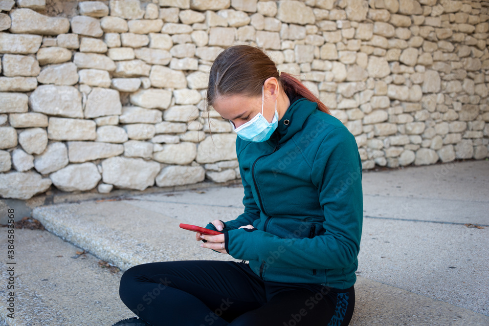 Fototapeta premium young sporty looking woman wearing a mask with mobile phone sitting outside ,covid 19 .