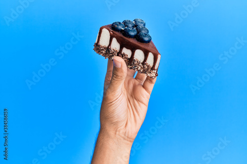 Hand of hispanic man holding slice of chocolate cake over isolated blue background.