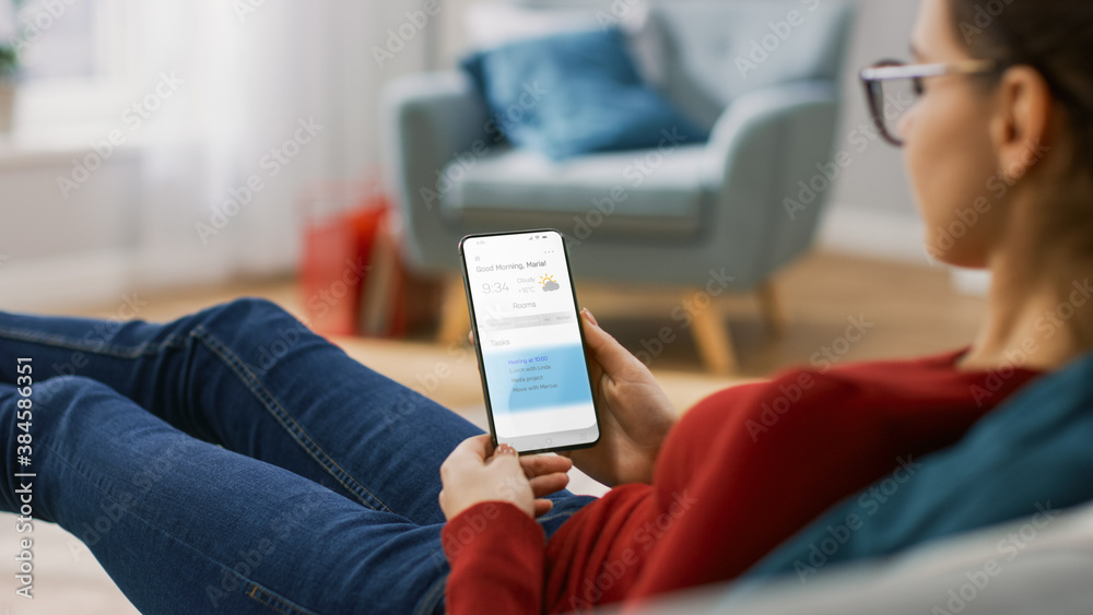 Young Woman at Home Uses Smartphone for Checking Weather Predictions and Coming Day Plans in Daily Schedule Calendar. She's Sitting On a Couch in Her Cozy Living Room. Over the Shoulder Shot Young Woman at Home Uses Smartphone for Checking Weather Predictions and Coming Day Plans in Daily Schedule Calendar. She's Sitting On a Couch in Her Cozy Living Room. Over the Shoulder Shot