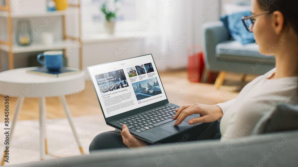 Young Woman at Home Is Using Laptop Computer for Scrolling and Reading News about Technological Breakthroughs. She's Sitting On a Couch in His Cozy Living Room. Over the Shoulder Shot Young Woman at Home Is Using Laptop Computer for Scrolling and Reading News about Technological Breakthroughs. She's Sitting On a Couch in His Cozy Living Room. Over the Shoulder Shot