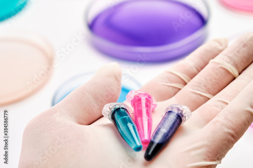 Eppendorf tubes with blue, green and pink liquids in the hand of scientist on the background of Petri dishes for microchemical analysis. 