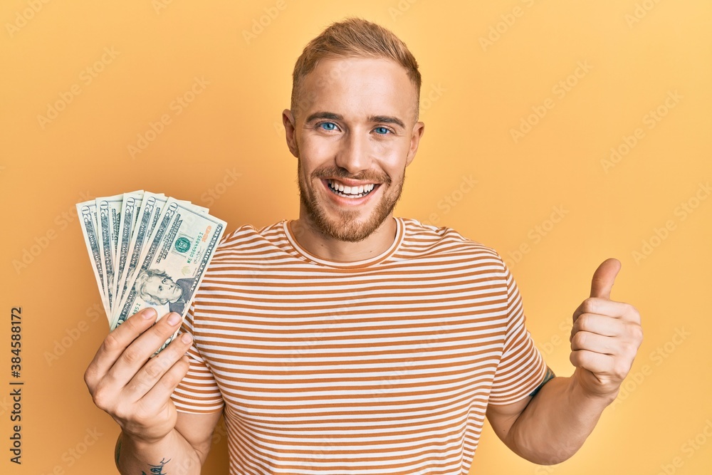 Young caucasian man holding dollars smiling happy and positive, thumb up doing excellent and approval sign Young caucasian man holding dollars smiling happy and positive, thumb up doing excellent and approval sign