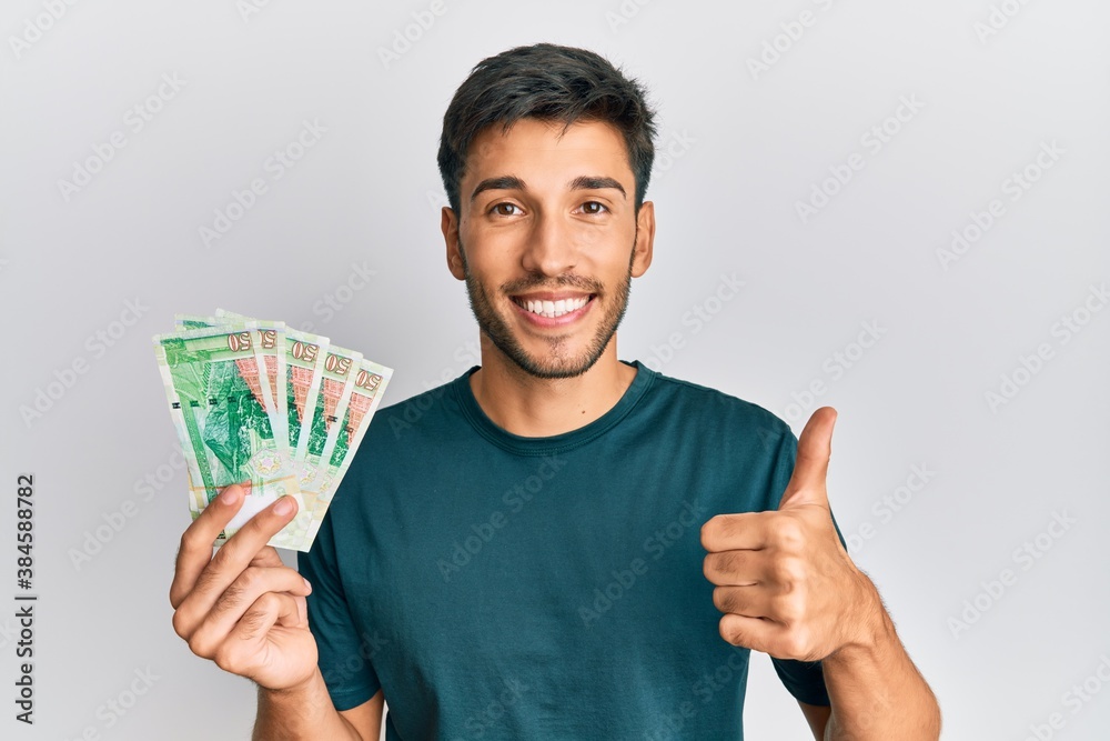 Young handsome man holding 50 hong kong dollars banknotes smiling happy and positive, thumb up doing excellent and approval sign Young handsome man holding 50 hong kong dollars banknotes smiling happy and positive, thumb up doing excellent and approval sign