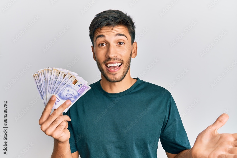 Young handsome man holding 20 swedish krona banknotes celebrating achievement with happy smile and winner expression with raised hand Young handsome man holding 20 swedish krona banknotes celebrating achievement with happy smile and winner expression with raised hand